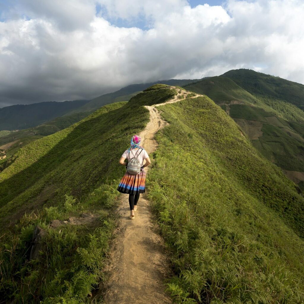 A lone hiker walking on a scenic mountain trail surrounded by lush greenery and clear blue skies.