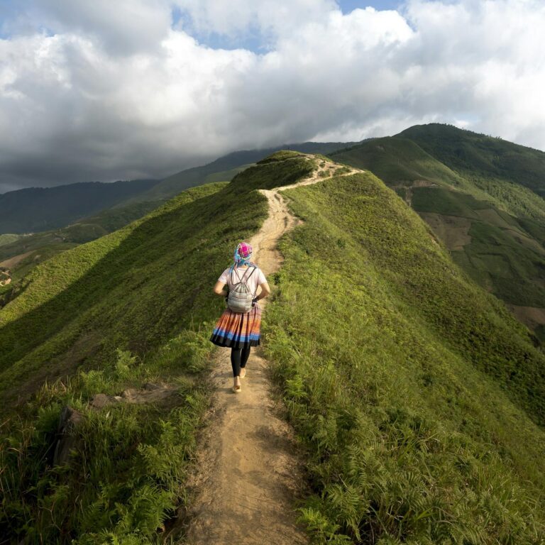 A lone hiker walking on a scenic mountain trail surrounded by lush greenery and clear blue skies.